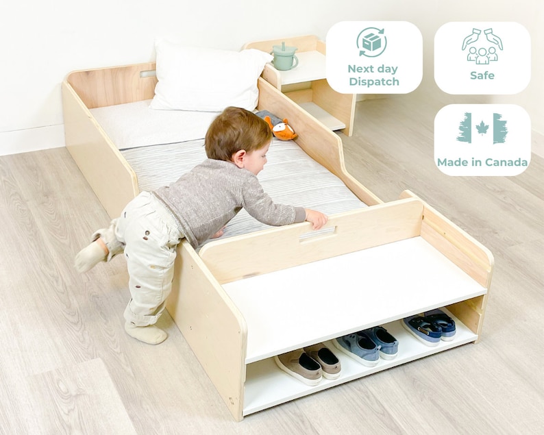 May include: A light wood toddler bed with a white mattress and a white shelf underneath. The shelf has a cutout for a handle and is holding several pairs of shoes. A young child is standing next to the bed, reaching for the handle.
