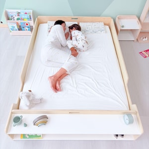 May include: A light wood-framed bed with a white mattress. A person and a child are sleeping on the bed. A shelf below the bed holds a toy, a small clock, and other items. A bookshelf and a nightstand are also visible.