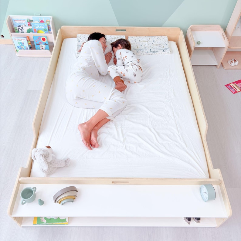 May include: A light wood-framed bed with a white mattress. A person and a child are sleeping on the bed. A shelf below the bed holds a toy, a small clock, and other items. A bookshelf and a nightstand are also visible.