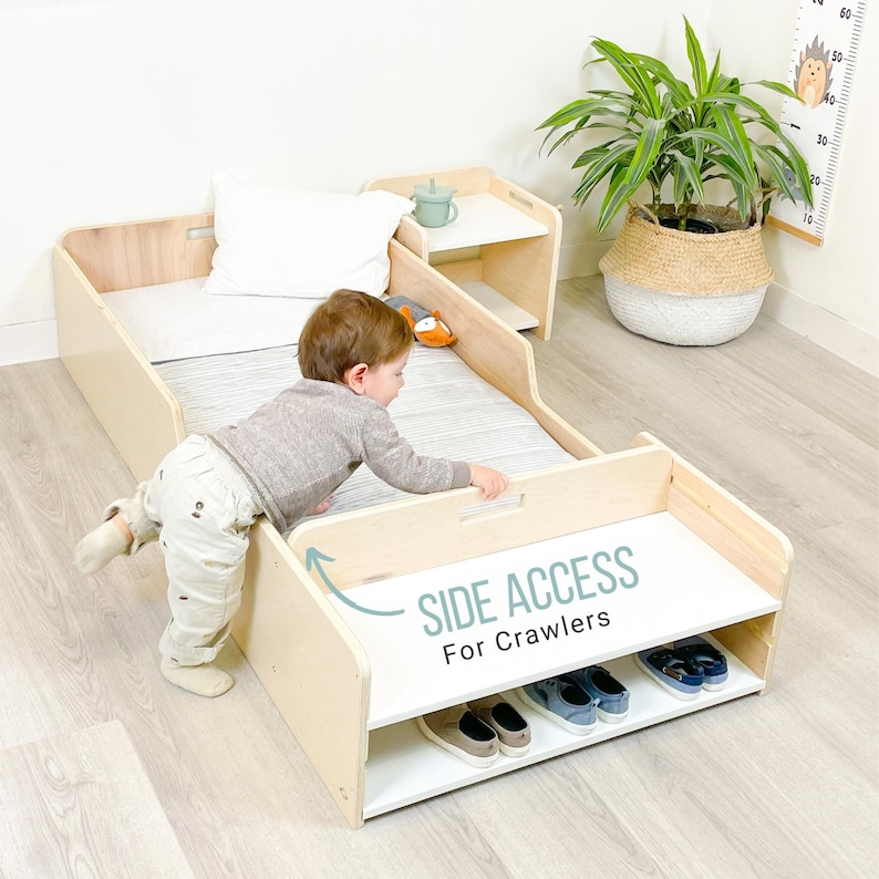 May include: A light wood toddler bed with a white mattress and a side access panel for crawlers. The bed has a built-in shelf for storage and a small side table with a cup holder. A child is crawling into the bed.