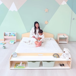 May include: A light wood bed frame with a white mattress, a white shelf with two mint green bins, and a small wooden nightstand. A woman and a child are sitting on the bed. The wall is painted with a mountain scene.
