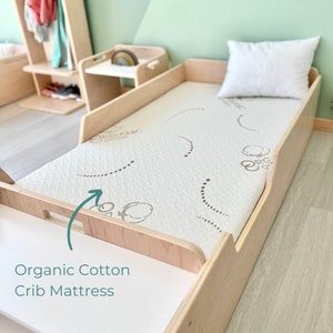 May include: A close-up of a light wood crib mattress with a white organic cotton mattress pad. The mattress pad has a subtle pattern of small circles and the words "Organic Cotton" printed on it. The mattress is in a crib with a light wood frame.
