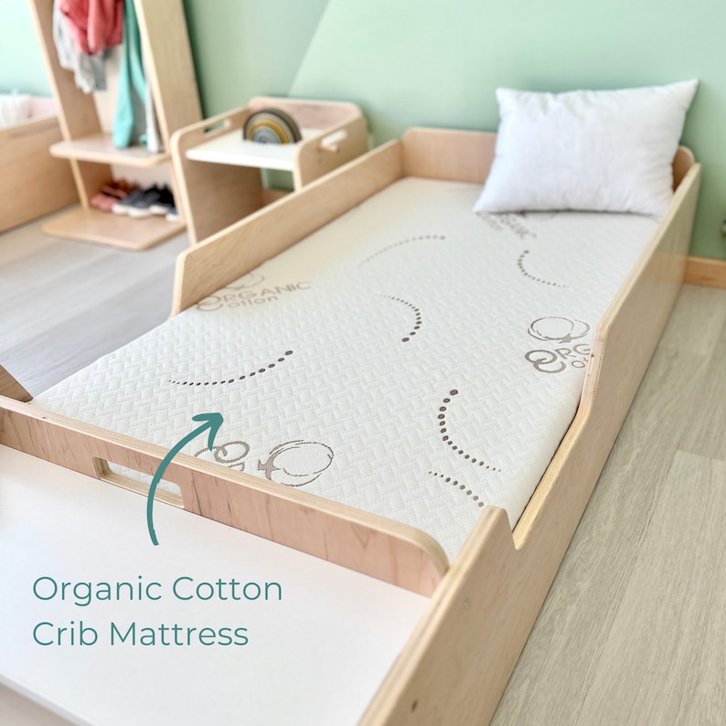 May include: A close-up of a light wood crib mattress with a white organic cotton mattress pad. The mattress pad has a subtle pattern of small circles and the words "Organic Cotton" printed on it. The mattress is in a crib with a light wood frame.