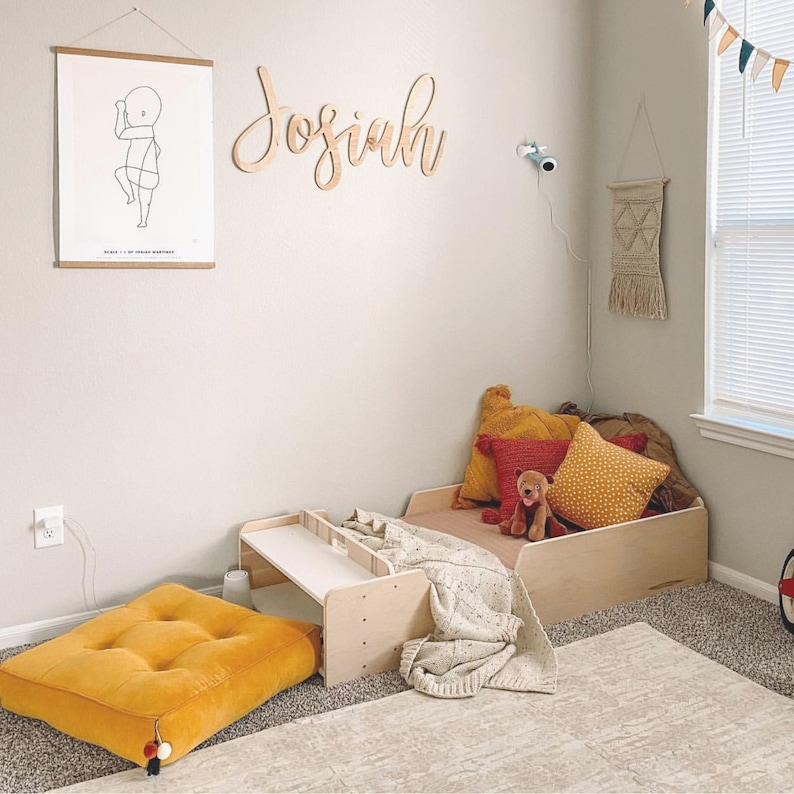 May include: A light wood bed frame with a white blanket and a stuffed animal. The bed is decorated with yellow and red cushions. The room has a white wall with a wooden name sign that says "Josiah".