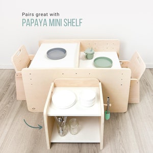 May include: A wooden play table with two chairs and a small shelf. The table has two white surfaces with a gray and green plate on each. The shelf has a white surface with two white plates, a glass jar, and a set of silverware. The shelf is labeled "Papaya Mini Shelf".