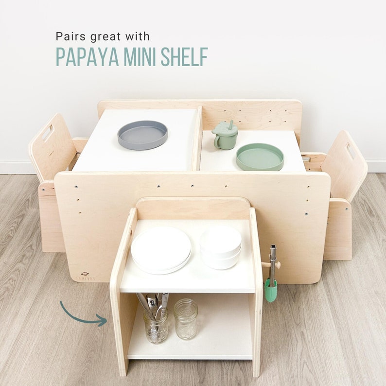 May include: A wooden play table with two chairs and a small shelf. The table has two white surfaces with a gray and green plate on each. The shelf has a white surface with two white plates, a glass jar, and a set of silverware. The shelf is labeled "Papaya Mini Shelf".