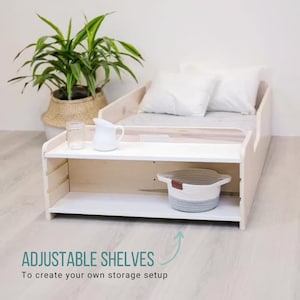 May include: A white wooden bed frame with adjustable shelves. The shelves are made of white wood and have a white finish. The bed frame is made of natural wood and has a light wood finish. The bed frame has a white mattress and two white pillows. The bed frame is sitting on a light wood floor. The text "ADJUSTABLE SHELVES" is written in green on a white background. The text "To create your own storage setup" is written in black on a white background.