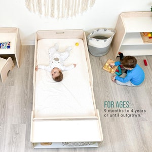 May include: A white toddler bed with a white mattress and a baby lying on it. The bed has a small shelf underneath. The text "FOR AGES: 9 months to 4 years or until outgrown." is visible in the image.