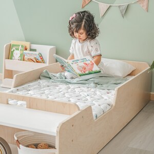 May include: A light wood toddler bed with a white mattress and a green and white patterned quilt. A young girl is sitting on the bed reading a book titled "Rocking Farm Animals".