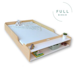 May include: A light wood changing table with a white changing pad. The table has a lower shelf with a green bin, a small plant, and a pair of brown shoes. A stuffed animal and a blanket are on the changing pad. The text "FULL BIRCH" is in the upper right corner.