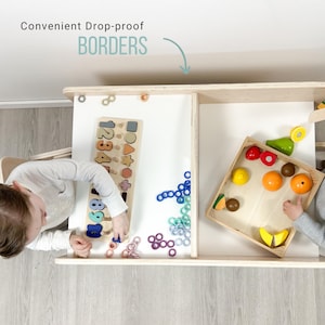 May include: Two children play at a wooden table with raised edges. One child is working on a number and shape puzzle, while the other is playing with a wooden crate filled with toy fruits and vegetables. The table has a white surface and is made of light wood.
