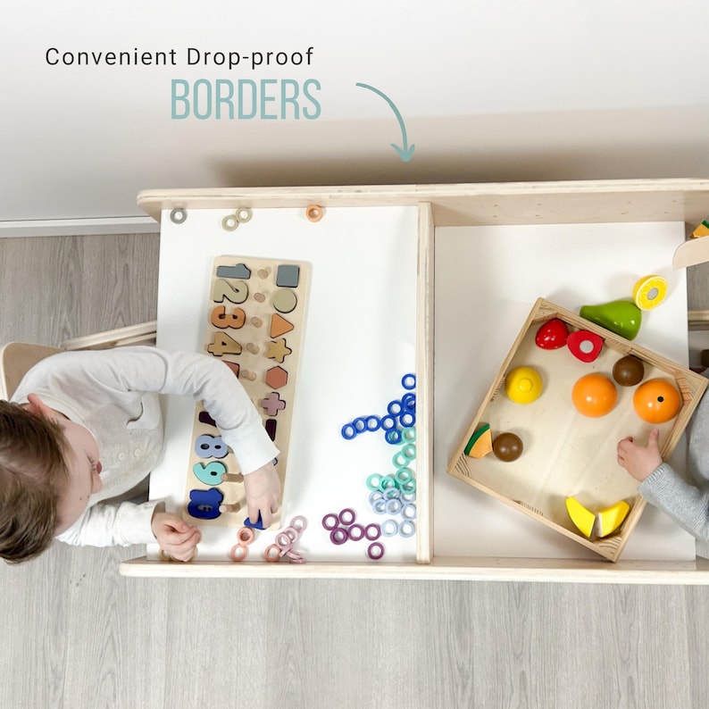 May include: Two children play at a wooden table with raised edges. One child is working on a number and shape puzzle, while the other is playing with a wooden crate filled with toy fruits and vegetables. The table has a white surface and is made of light wood.
