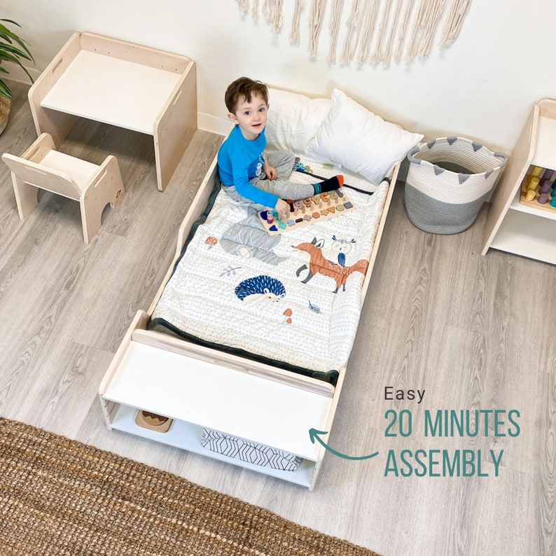 May include: A white wooden toddler bed with a white mattress and a colourful quilt. A child is sitting on the bed playing with a wooden toy. The bed has a shelf underneath. Easy 20 Minutes Assembly.