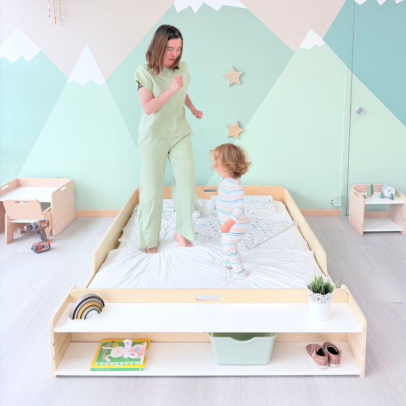 May include: A light wood-framed toddler bed with a white mattress and a lower shelf. The shelf holds a rainbow, a book, a green bin, and shoes. A woman and a toddler are on the bed. The wall is painted with a mountain scene.