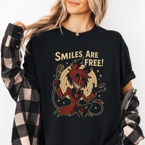 May include: Black t-shirt featuring a cartoon character with a microphone, wearing a red suit and a wide grin. The text "SMILES ARE FREE!" is above the character. The design is set against a beige background with star accents.