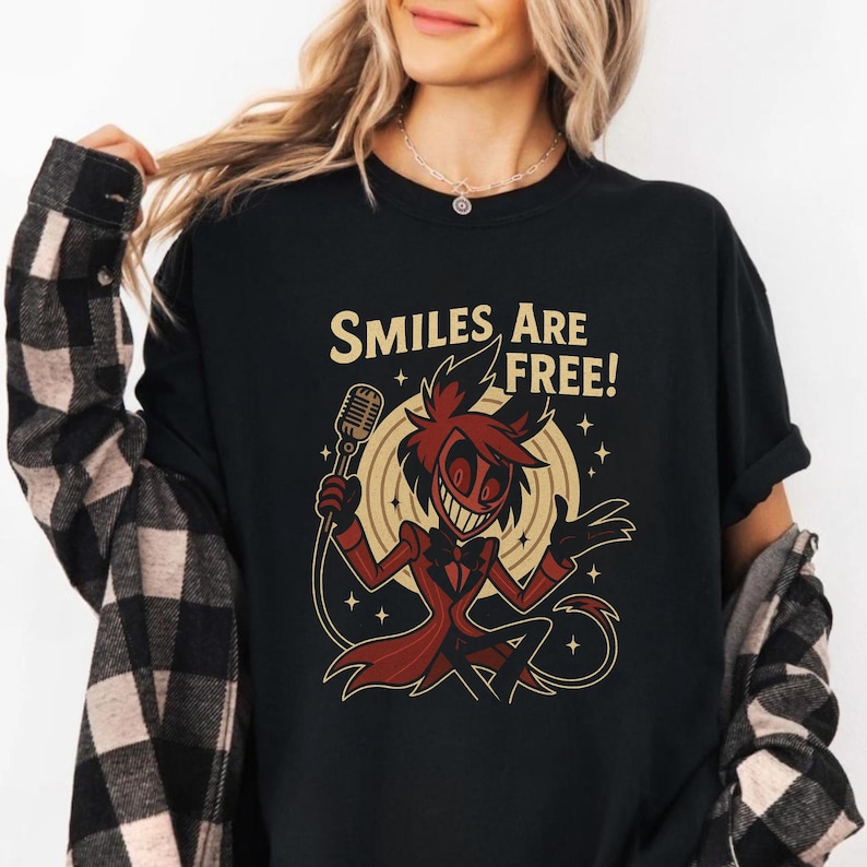 May include: Black t-shirt featuring a cartoon character with a microphone, wearing a red suit and a wide grin. The text "SMILES ARE FREE!" is above the character. The design is set against a beige background with star accents.