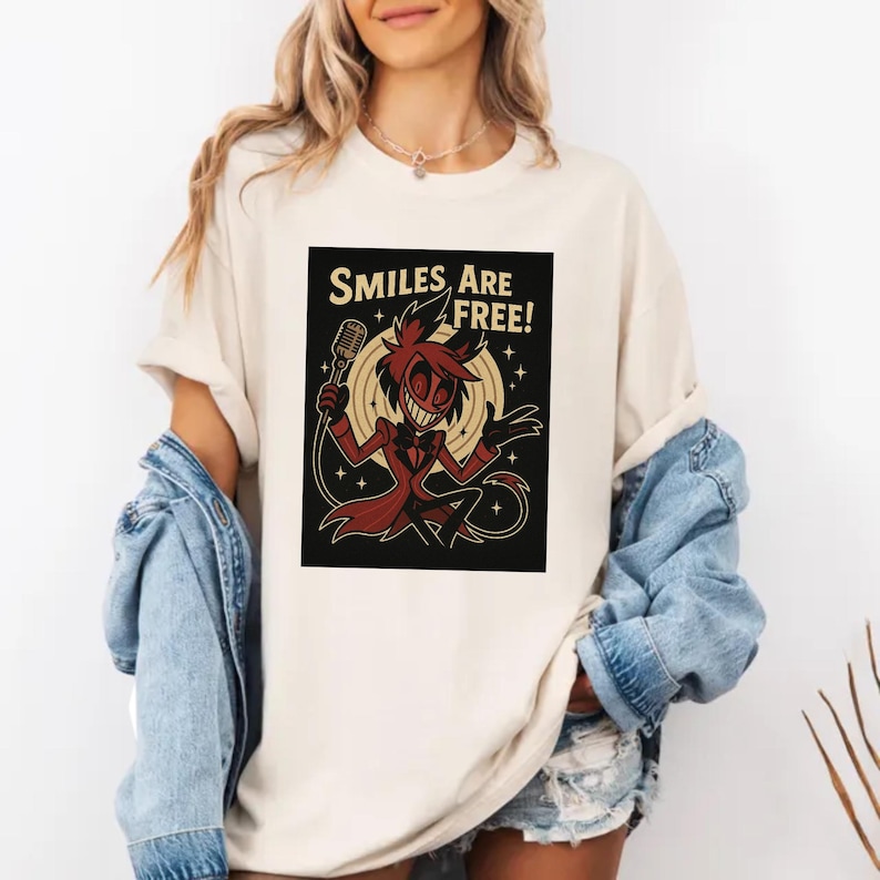 May include: Cream-colored t-shirt featuring a cartoon character holding a microphone, with the text "SMILES ARE FREE!". The character is depicted in red and black, set against a black background with a yellow circle and stars.