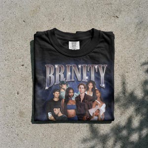 Può includere: Una t-shirt nera a girocollo con una stampa grafica che riporta la parola BRINITY in un carattere argento metallizzato. Il design include un collage di sei persone in un'ambientazione scura e suggestiva con un'etichetta Comfort Colors.