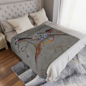 May include: A gray blanket featuring a Native American-inspired design. The artwork depicts a figure on horseback with a bow and arrow, surrounded by birds and abstract symbols. The blanket is draped over a bed with white bedding.