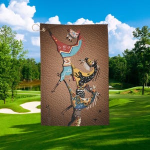 May include: A golf towel featuring a stylized illustration of Native American figures in shades of red, blue, yellow, and brown. The design is set against a textured brown background, with a gold-colored grommet for hanging.