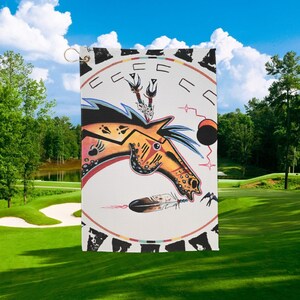 May include: A rectangular white golf flag featuring a Native American horse design. The horse is orange and yellow with black and red accents. The flag has a gold grommet for hanging and is set against a green golf course.