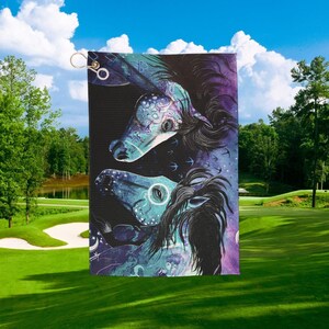 May include: Golf towel featuring a detailed illustration of three stylized horse heads in shades of teal, purple, and black. The design includes intricate patterns and a flowing mane. A gold-colored clip is attached to the top.