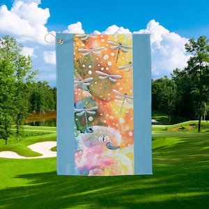 May include: A light blue golf towel with a dragonfly design. The center panel features a colorful illustration of dragonflies and a whimsical creature. The towel has a metal grommet for hanging and is set against a golf course backdrop.