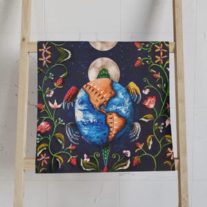 May include: A decorative towel featuring a vibrant illustration of the Earth with wings, surrounded by floral and celestial elements. The artwork includes a blue and brown globe, red and yellow wings, and a dark blue background with flowers and moons.