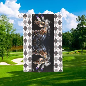 May include: A golf towel featuring a detailed illustration of two horses with Native American-inspired designs. The horses are set against a black background, framed by a gray, white, and red argyle pattern. The towel has a gold grommet for hanging.