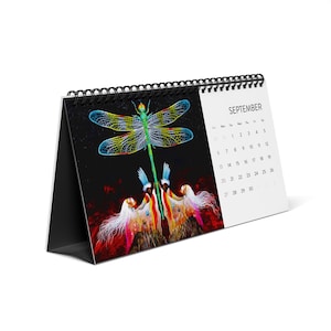 May include: A desk calendar featuring a vibrant dragonfly illustration with a black background. The dragonfly is green, yellow, and blue. The calendar displays the month of September with dates. The calendar is held together with a black spiral binding.