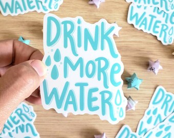 May include: A white sticker with blue text that reads "Drink More Water". The sticker has a whimsical, hand-drawn style with water droplets around the text.