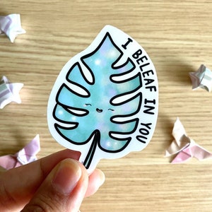 May include: A white sticker with a blue and green monstera leaf with a smiling face. The text "I Beleaf in You" is written on the sticker.