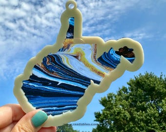 May include: A cream-colored West Virginia state outline pendant with a marbled blue, gold, and white design. The pendant has a small hole at the top for hanging. The background shows a blue sky and green trees.