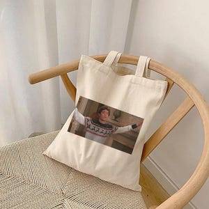 Puede incluir: Una bolsa de lona blanca con una imagen impresa de una persona que lleva un suéter estampado y extiende los brazos. La persona está de pie frente a una chimenea.