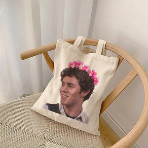 Puede incluir: Bolso tote de color crema con la imagen de una persona sonriente con una corona de corazones rosas. El bolso tiene dos asas y está sobre una silla de madera.