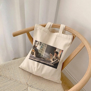 Puede incluir: Una bolsa de lona blanca con una imagen impresa de una persona sentada en una cama, leyendo un libro. La bolsa tiene dos asas.