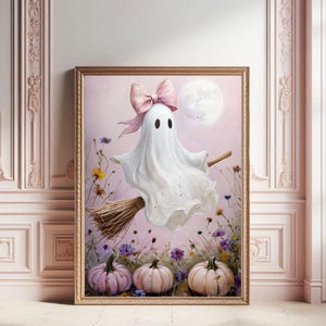May include: A framed painting depicts a white ghost with a pink bow, flying on a broom against a pink sky with a full moon. The artwork includes pumpkins and wildflowers, creating a charming Halloween theme.