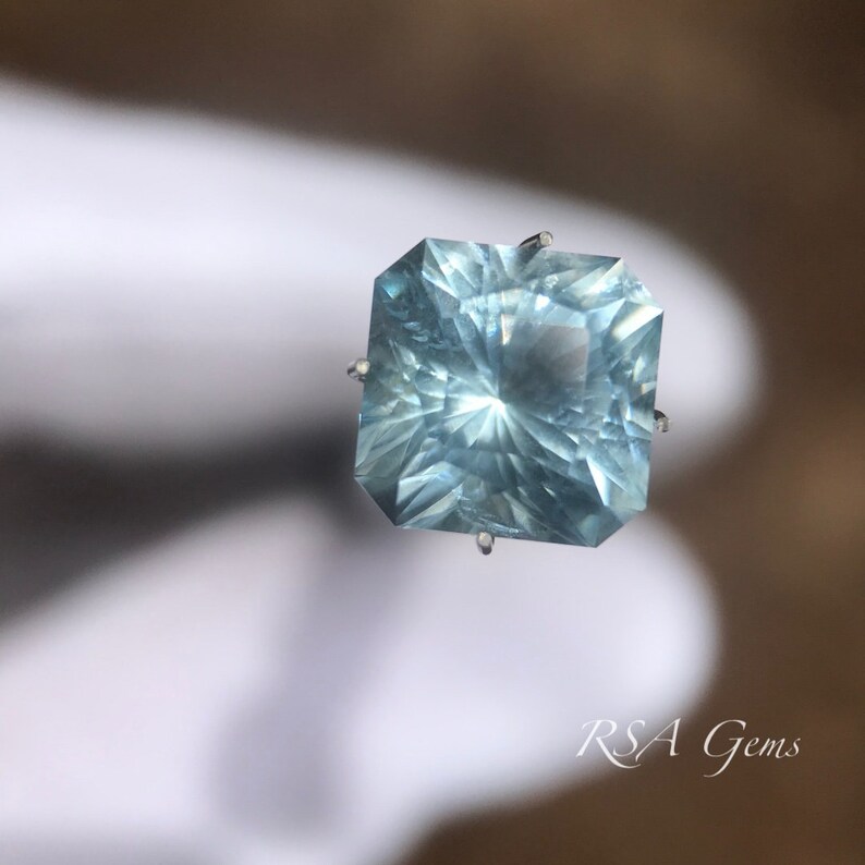 May include: A light blue aquamarine gemstone with a square shape and a faceted cut. The gemstone is set in a silver prong setting.