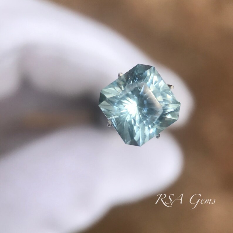 May include: A light blue aquamarine gemstone with a square cut and a faceted design. The gemstone is set in a silver prong setting.
