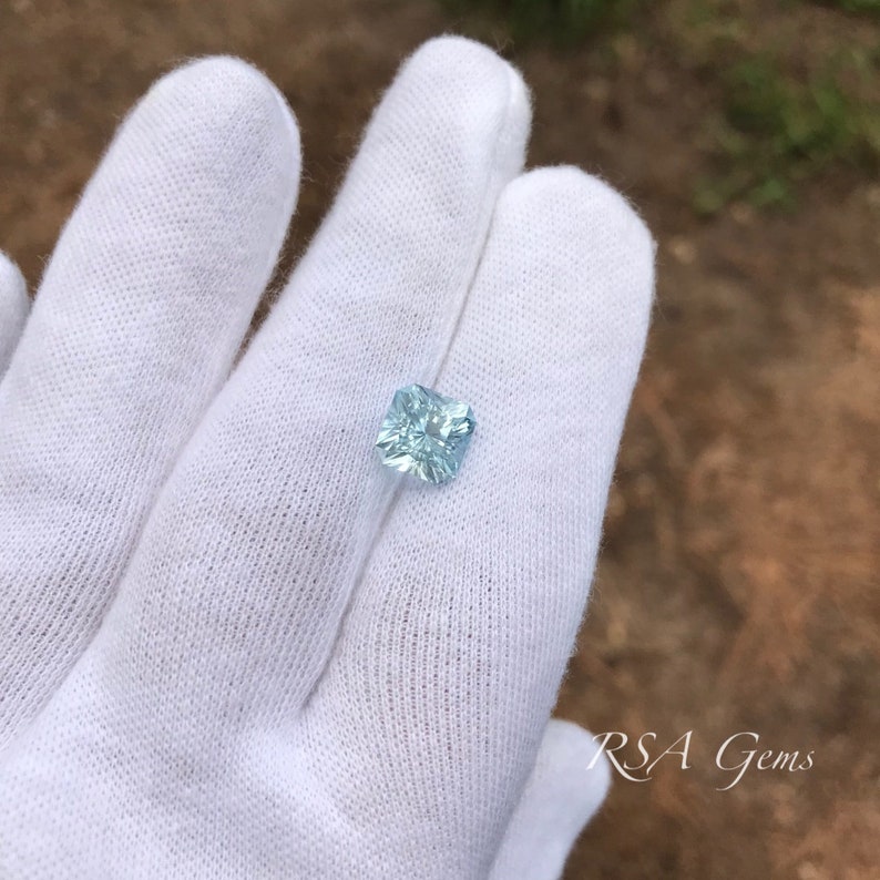 May include: A light blue square-shaped gemstone with facets, held in a white gloved hand.  RSA Gems is printed in the bottom right corner of the image.