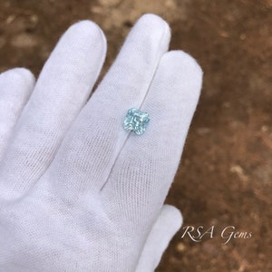 May include: A light blue square-shaped gemstone with facets, held in a white gloved hand. The gemstone is a light blue colour and has a slight sparkle. The background is a brown, textured surface.  RSA Gems is printed in black text on the bottom right corner of the image.