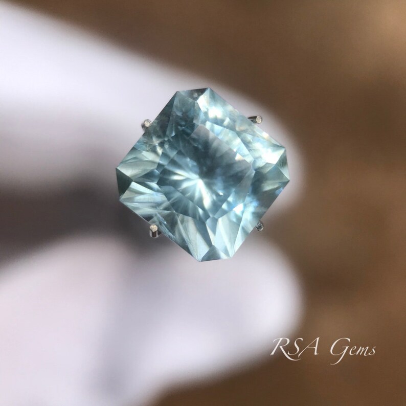May include: A light blue aquamarine gemstone with a square shape and many facets. The gemstone is set on a clear stand.