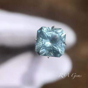 May include: A light blue gemstone with a square cut and a faceted design. The gemstone is held in place by four prongs.
