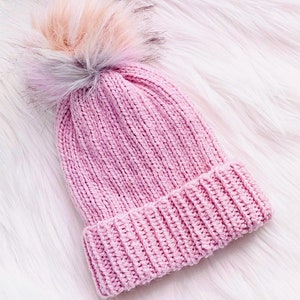 May include: A pink knitted beanie with a fluffy multi-coloured pom pom on top. The beanie is on a white fluffy surface.