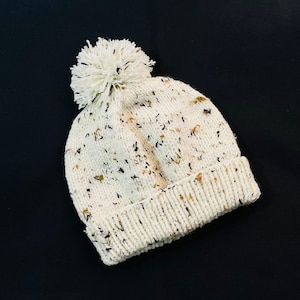 May include: A white knitted beanie with a fluffy pom pom on top. The beanie has a speckled pattern with brown and black flecks.