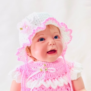 May include: A pink crocheted dress with white trim and a matching white crocheted hat with pink trim. The hat has a small flower embellishment.