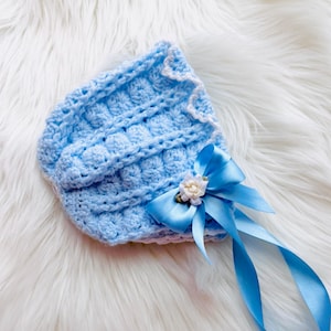 May include: A light blue crocheted bonnet featuring a textured bobble stitch pattern, a scalloped white trim along the edge, and a decorative blue satin ribbon bow accented with a small white fabric flower and green leaves at the side.