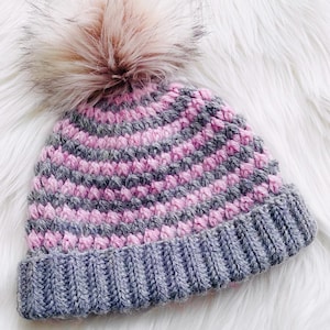 May include: A gray and pink striped crocheted beanie with a fluffy pom pom on top.