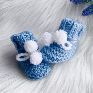 May include: A pair of handmade baby booties in a light blue knit. Each bootie features a white pom-pom and a delicate white tie. The booties are displayed on a soft, white, faux fur surface, with blue flowers in the background.