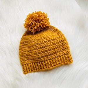 Puede incluir: Gorro tejido de color amarillo mostaza con un pompón a juego en la parte superior. El gorro está hecho de una tela de punto texturizada y se muestra sobre una superficie blanca y esponjosa. Un gorro cálido para el clima frío.